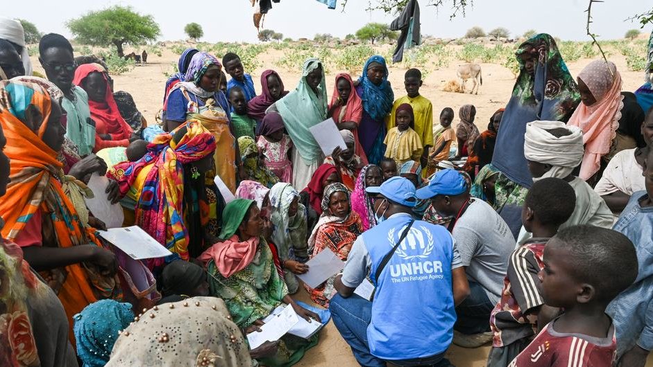 Wars that outlast attention: The role of humanitarian diplomacy 1 The image shows a photograph of a group of people huddled around a man in a UNHCR T-shirt